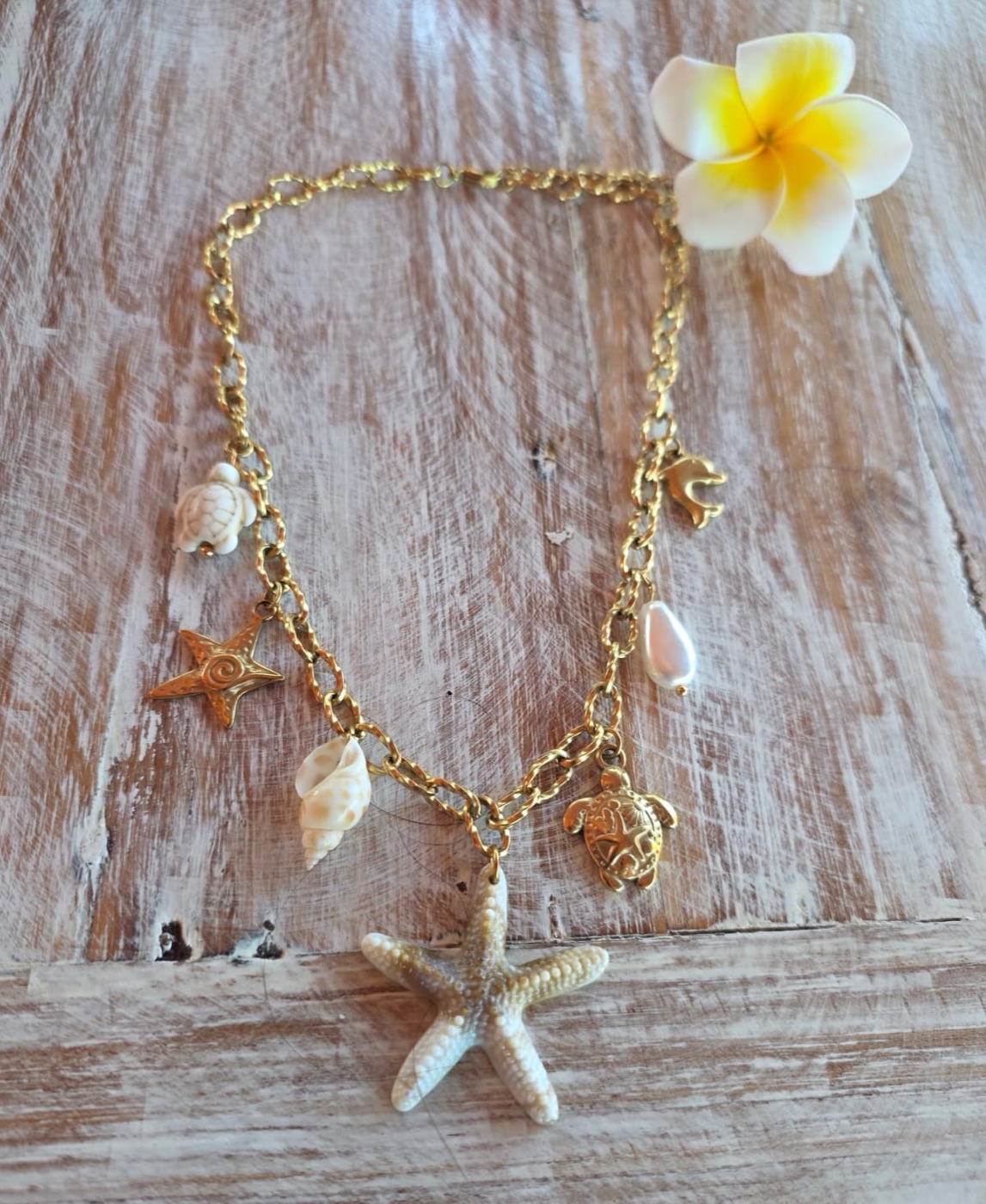 Gold chain necklace hung with starfish, shells and turtle charms, on weathered wood beside a frangipani flower